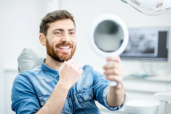 Bearded white patient checking out his handsome smile in a mirror while sitting in a dental chair.